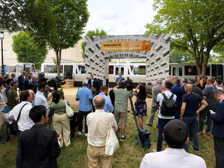 A crowd gathers at a Metro event to celebrate the 50th anniversary of the Washington DC Metro system, with speakers at a podium and media coverage. The backdrop features the text "welcome to america's metro system" and a modern design, while lush trees provide shade in the outdoor setting.