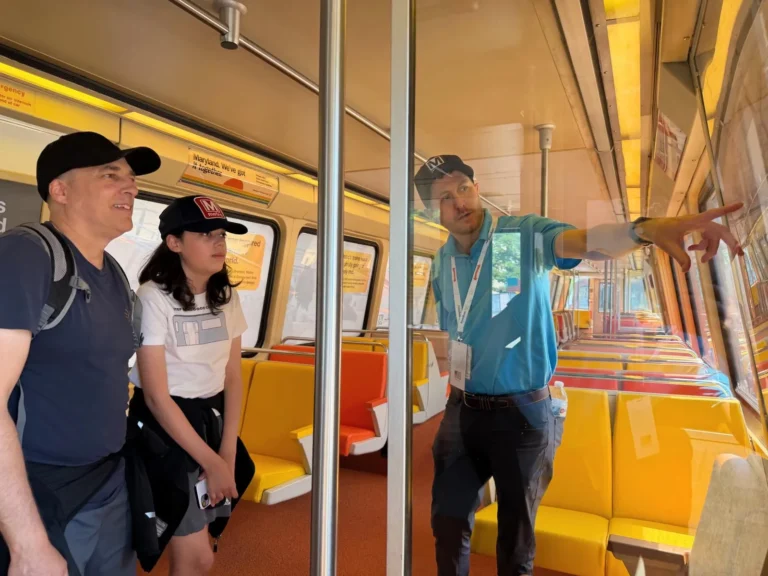 A metro employee points out details on a display to two visitors inside a colorful train car, celebrating the Washington DC Metro's 50th anniversary. The interior features bright orange and yellow benches, and informational posters are visible on the walls. The atmosphere reflects an educational and celebratory experience.