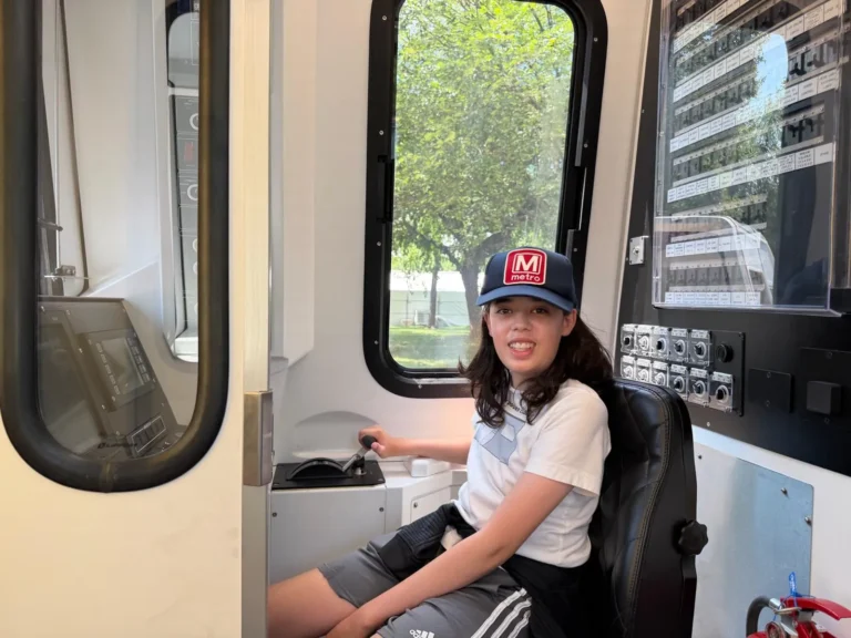 A young woman wearing a baseball cap sits in the driver's seat of a Metro train, smiling. The interior features control panels and a view of green trees outside, celebrating the Metro system's 50th anniversary in the Washington DC region.