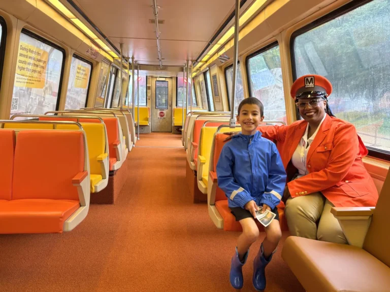 A smiling woman in a Metro uniform sits beside a young boy in a blue rain jacket and boots inside a nearly empty Metro train car, which features orange and yellow seats. The interior shows a cozy yet vibrant design, celebrating the 50th anniversary of the Washington DC Metro system.