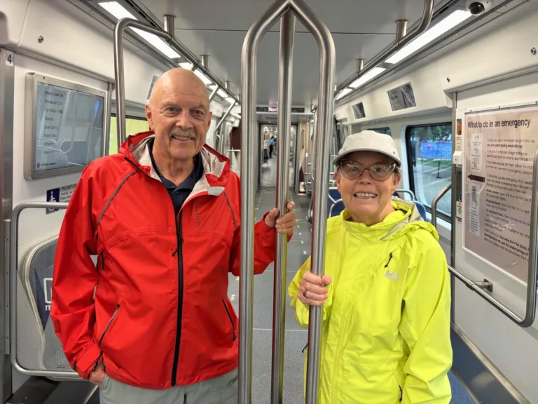 Two smiling seniors stand inside a clean, modern Metro train. One wears a red jacket and the other a bright yellow raincoat, both holding onto metal poles. The train is empty, showcasing the spacious interior with informational signs on the walls, highlighting the Metro's 50th anniversary in Washington, DC.