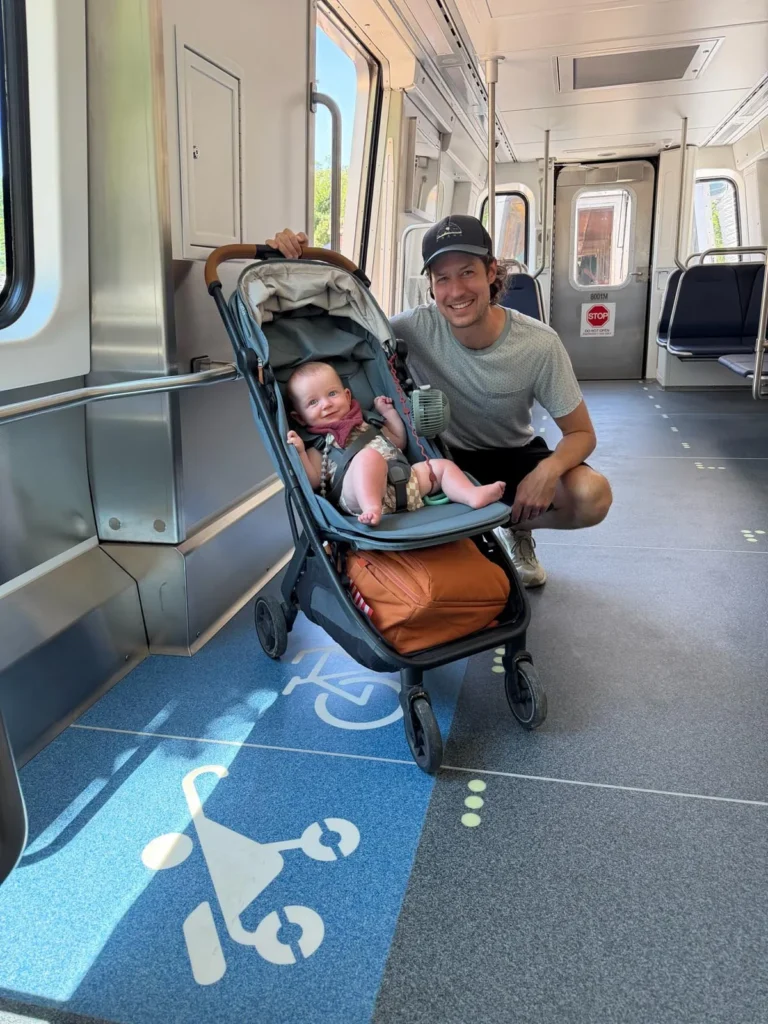 A man smiles while posing next to a stroller with a baby inside on a Metro train, highlighting family-friendly accessibility in the Washington DC Metro system, celebrating its 50th anniversary. The train's interior features modern design and signage promoting stroller use.