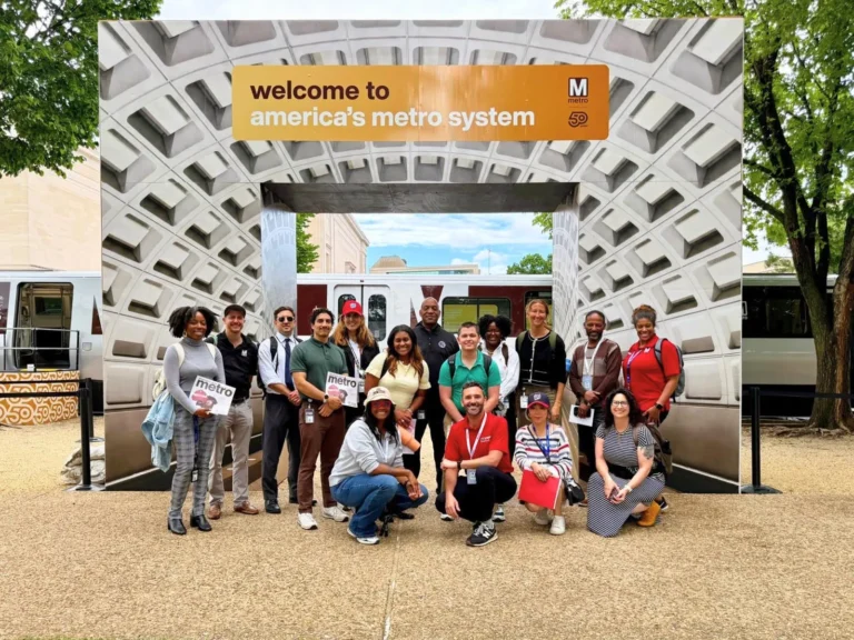 A diverse group of people poses together under a large welcome sign celebrating the 50th anniversary of America's Metro system, with a backdrop featuring a Metro train and distinctive architectural elements. The participants are smiling and holding Metro-themed materials, surrounded by greenery.