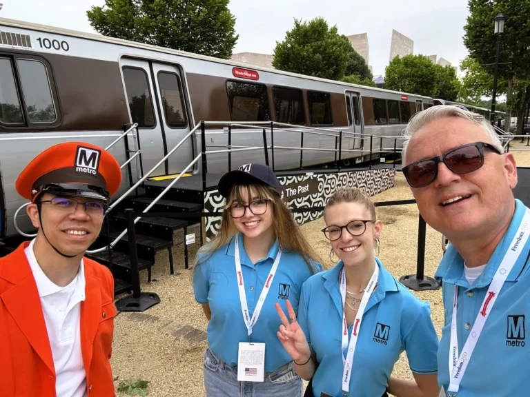 Four people pose in front of a Washington DC Metro train, celebrating its 50th anniversary. The group includes a man in an orange cap and uniform, two women in blue Metro t-shirts, and a man taking the selfie. They stand outside on a gravel path, with the train prominently displayed in the background.