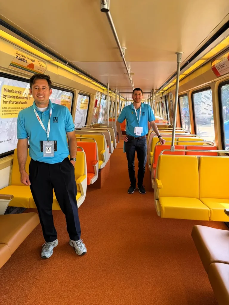 Two men in blue polos with Metro logos stand in a nearly empty subway car featuring bright orange and yellow seating, celebrating the 50th anniversary of the Washington DC Metro system. The interior has large windows allowing light in, highlighting the clean and modern design.
